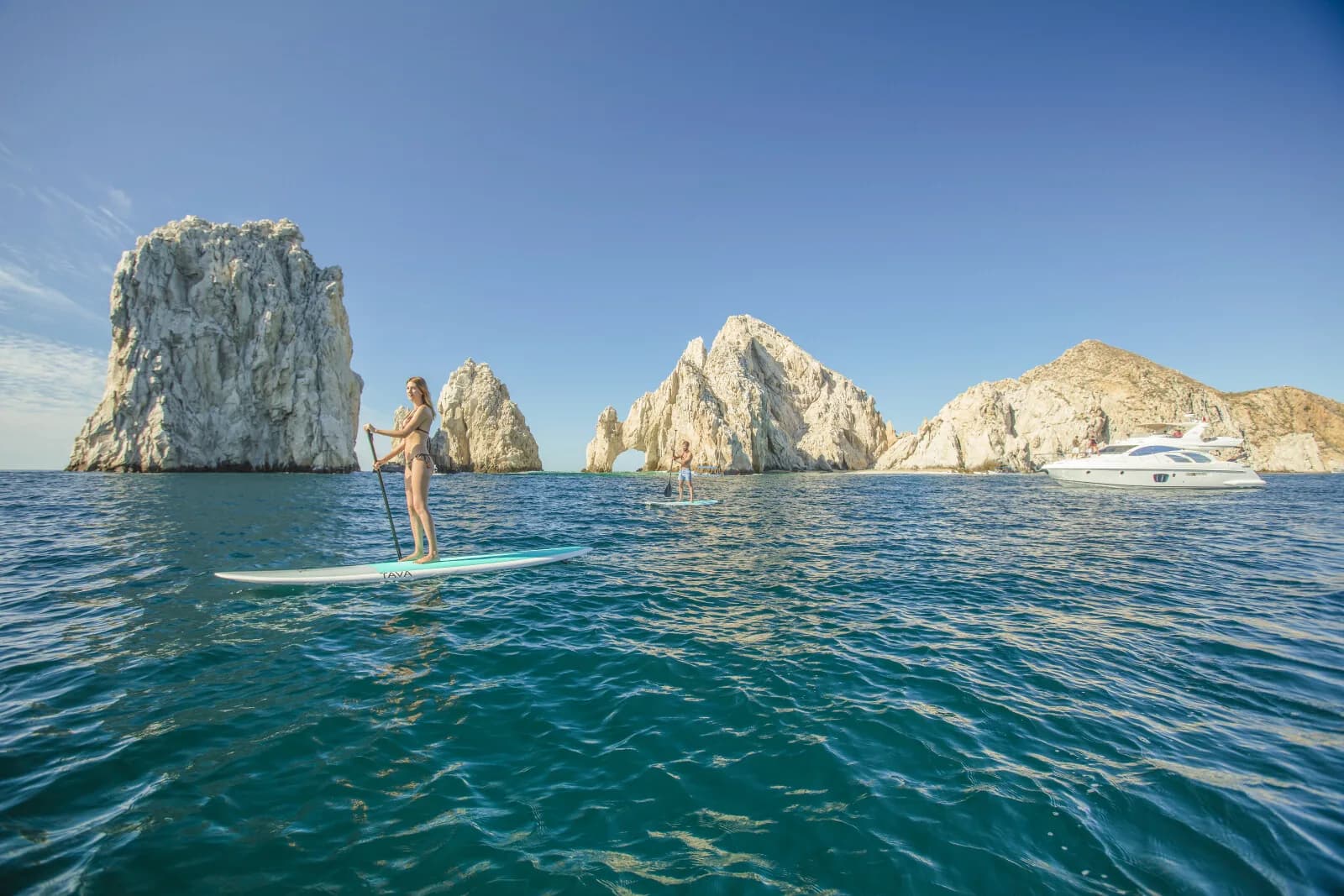 A companion paddleboarding on calm turquoise water off the coast.