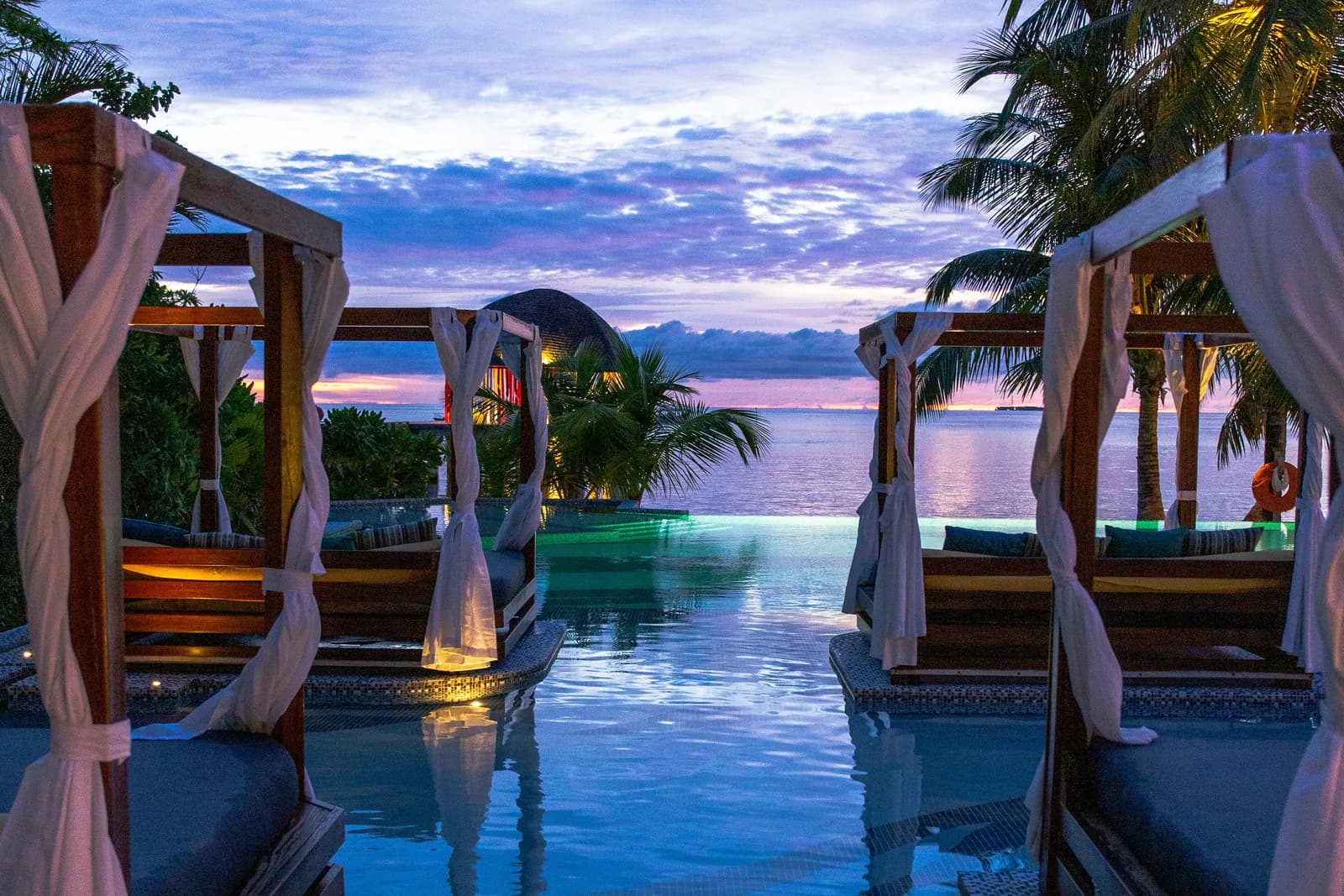Curtained cabanas beside an infinity pool looking out to the sea at dusk.