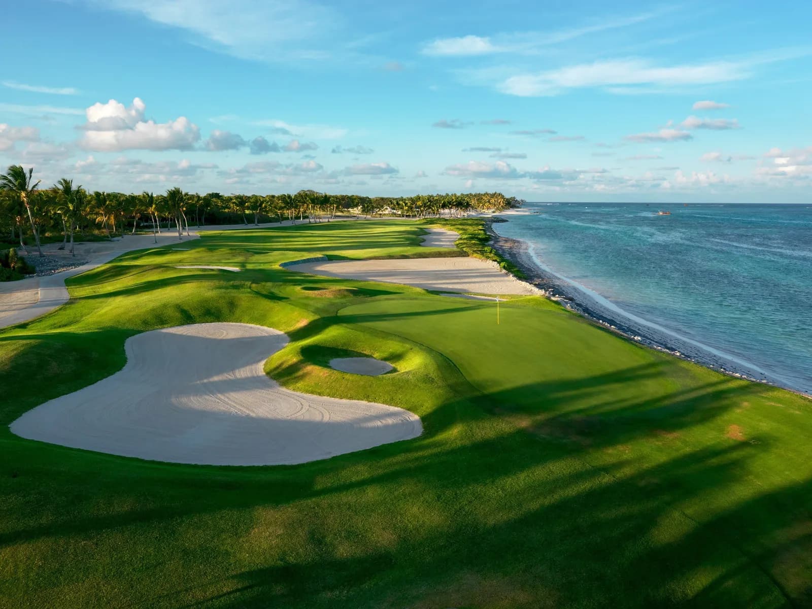 A La Cana green with its yellow flag beside the beach, a broad white bunker in front and turquoise Caribbean shallows running the length of the hole.