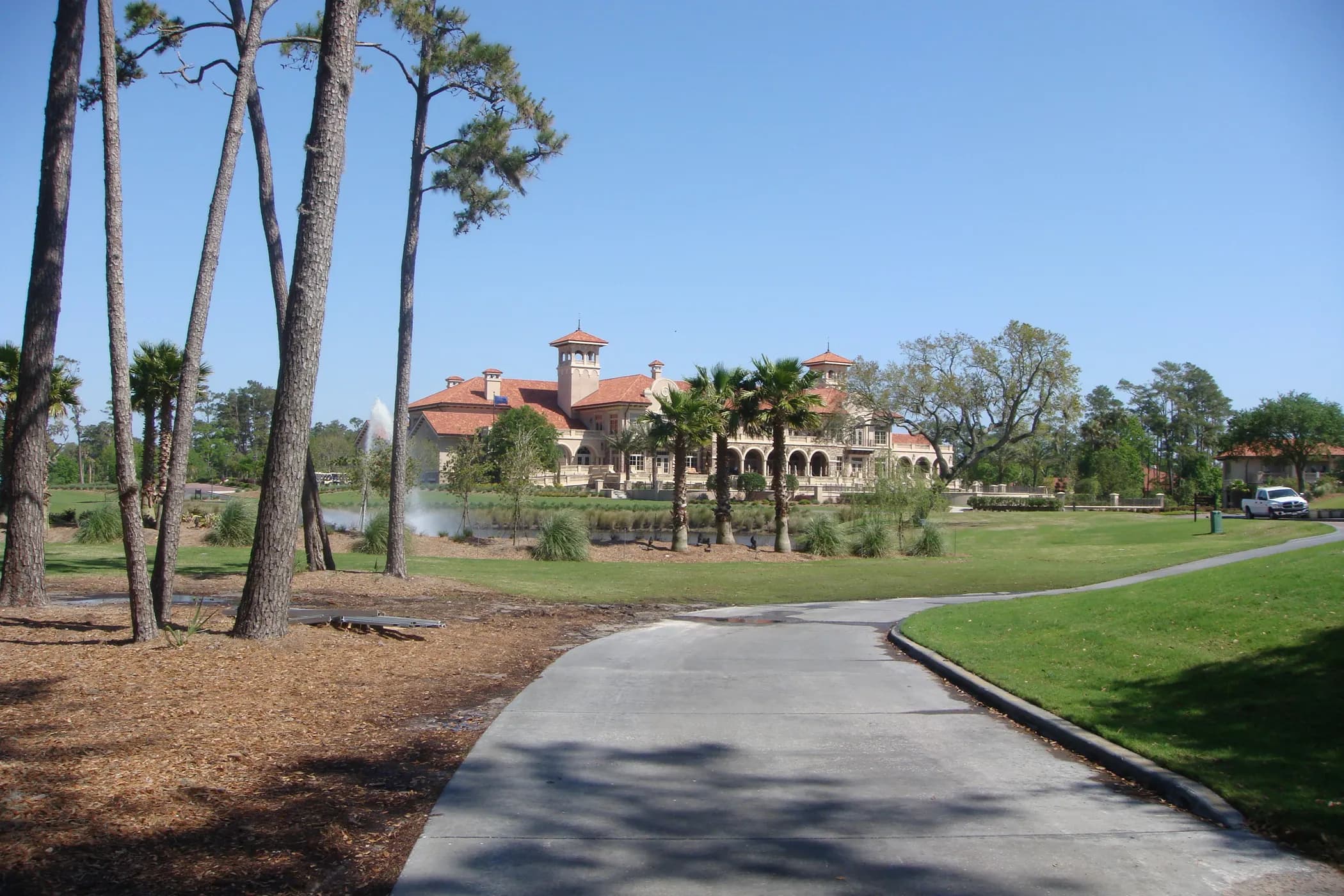 The Mediterranean-style TPC Sawgrass clubhouse beyond palms and a fountain pond, under a clear sky.