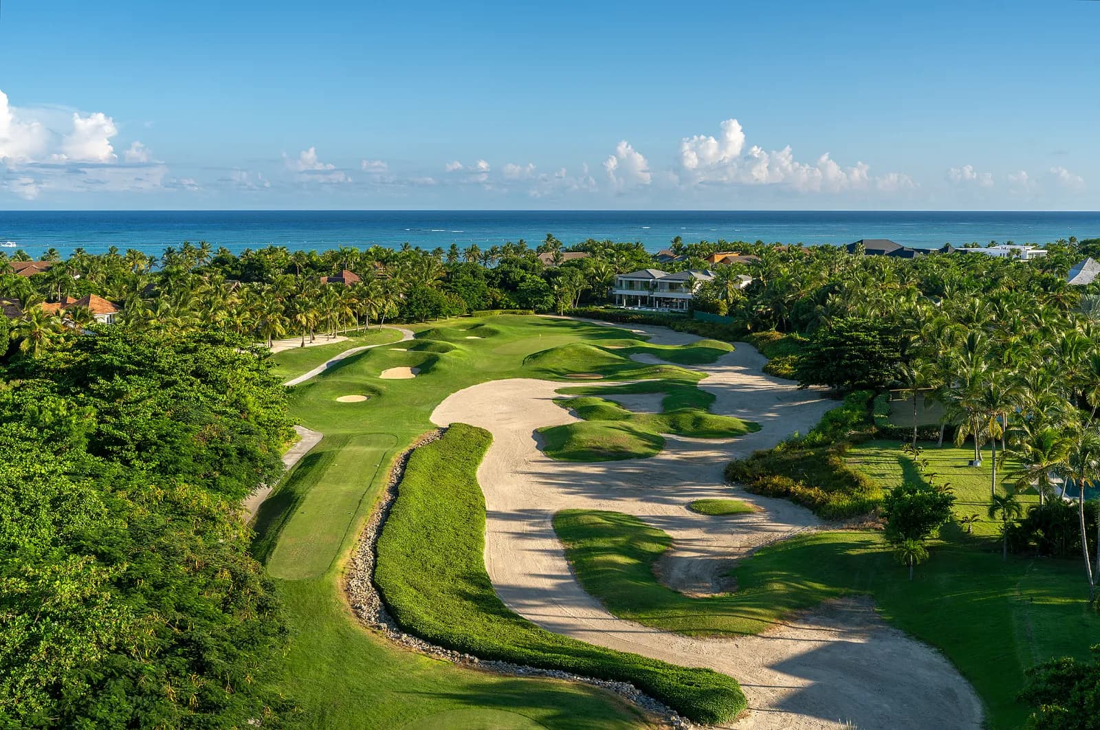 A fairway at La Cana Golf Club with a large sweeping sand bunker, palms and villas alongside, and the Caribbean Sea beyond the green.