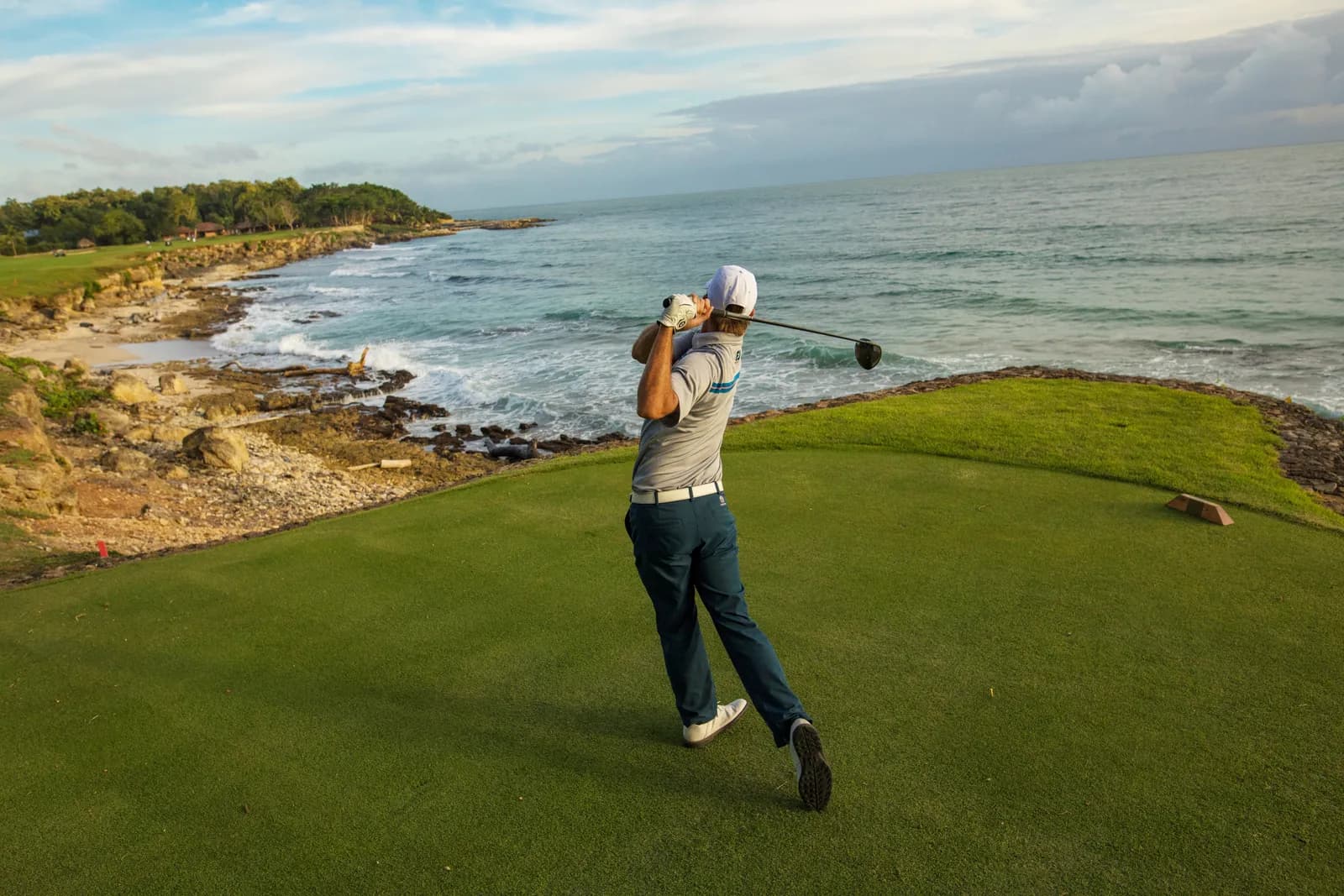 A golfer follows through from a seaside tee above a rocky cove.