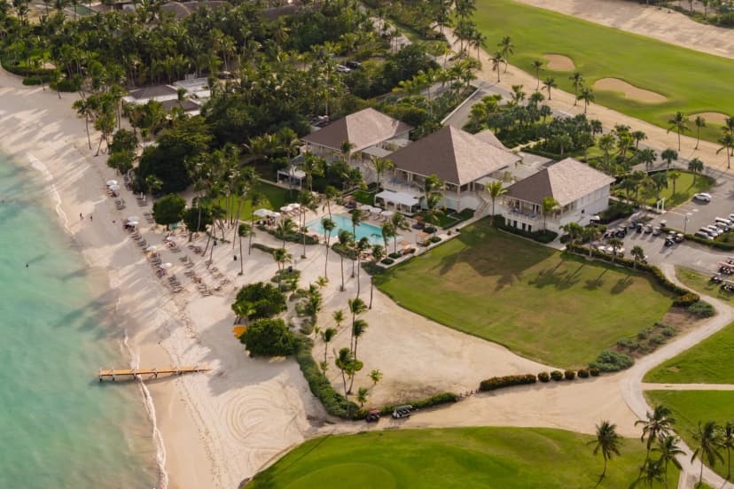 Aerial of the La Cana clubhouse and pool beside a white sand beach and turquoise water, with a pier and fairways alongside