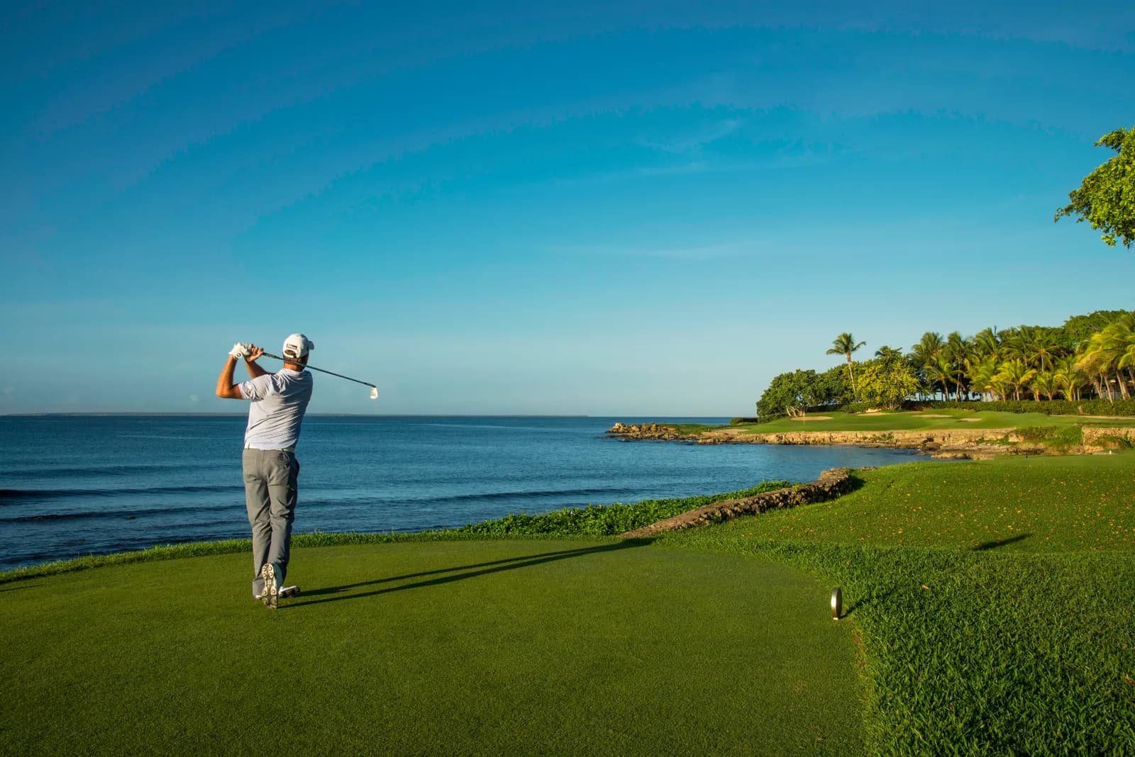 A golfer follows through on a seaside tee, hitting toward a green backed by palms across the water.