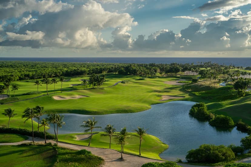 Aerial view over Corales Golf Course with wide fairways, bunkers, palm-lined lakes and the Caribbean Sea beyond, under towering clouds.