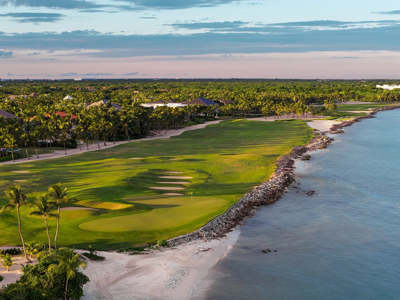 Aerial view of a seaside par 5 at La Cana, its green ringed by bunkers on a rocky point, palms and villas inland and a pale beach curving away.