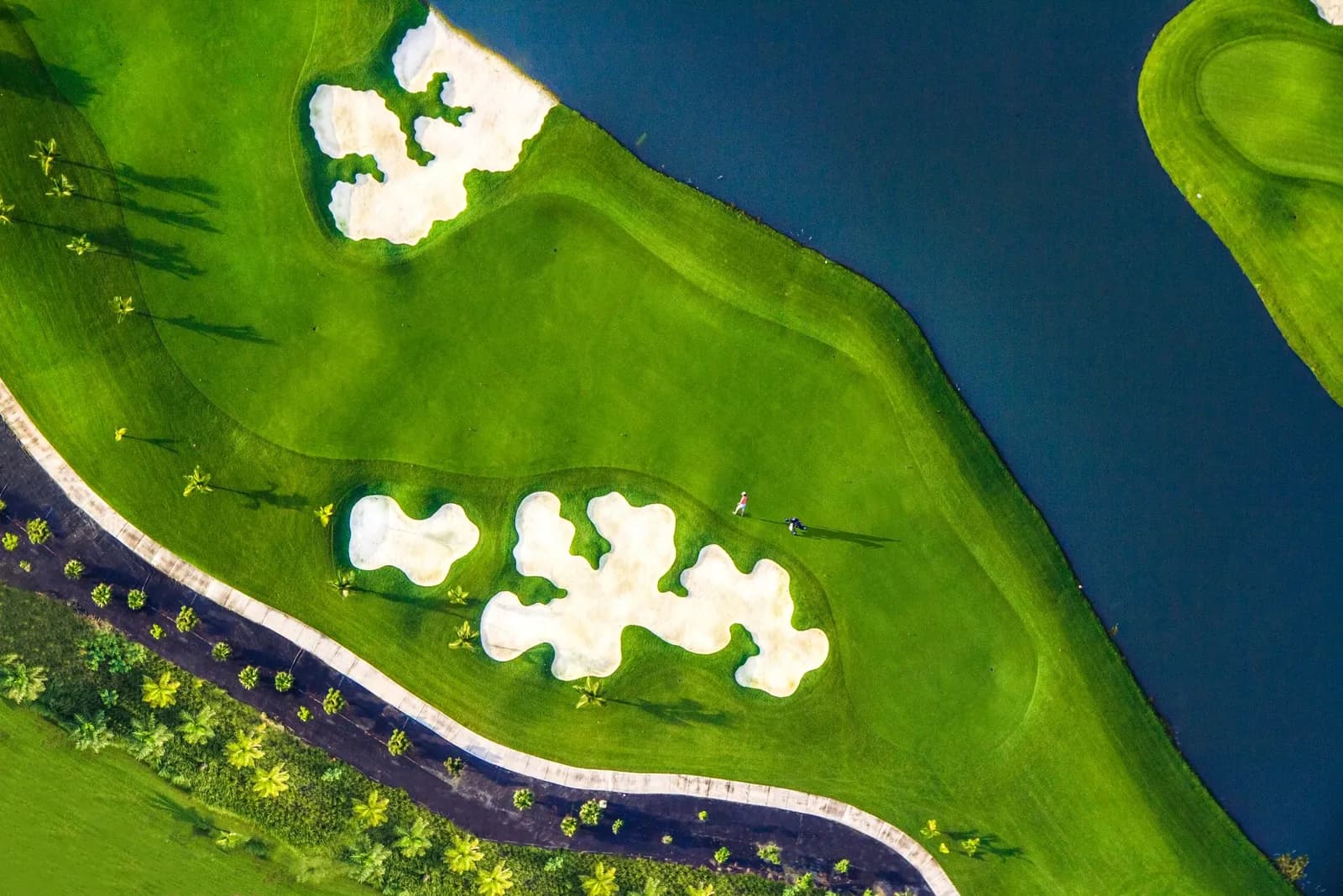 Top-down aerial of Santa Maria Golf Club: a green ringed by white cloverleaf bunkers beside a dark lake, a lone golfer and trolley on the fairway.