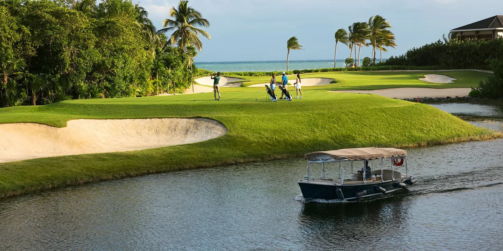 Golfers on an oceanfront green at El Camaleón as a boat passes on the canal