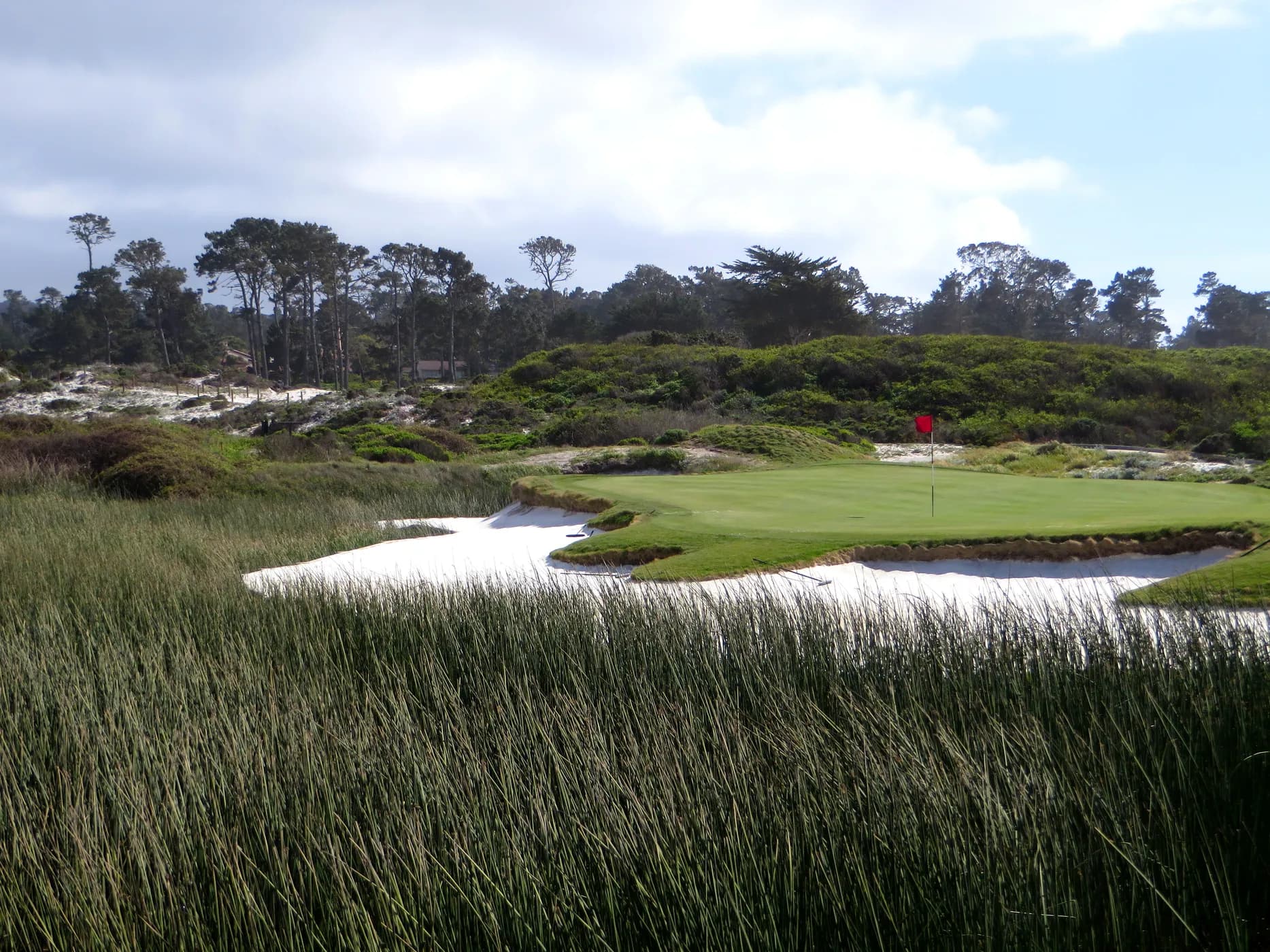 A green at The Links at Spanish Bay ringed by white sand bunkers and dune grasses, with Monterey pines on the ridge behind.
