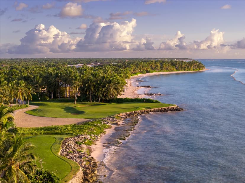An oceanfront green at La Cana Golf Club at sunset, set on a rocky shoreline beside a curving palm-lined beach.