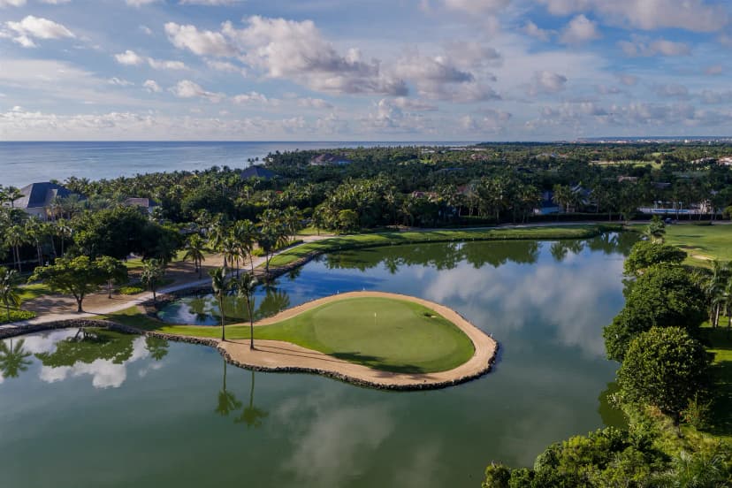 A green at La Cana Golf Club almost surrounded by a lake, ringed with palm trees, with villas and the Caribbean Sea behind.