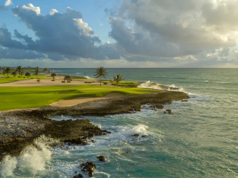 A Punta Espada green beside crashing surf on dark coral rock at golden hour, with fairway and palms behind