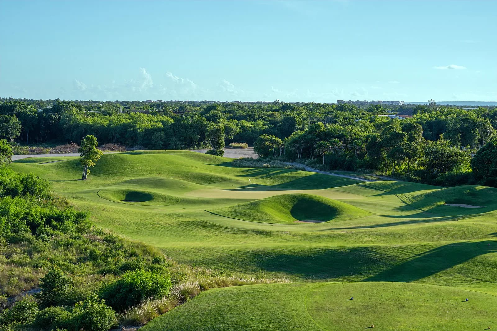 Elevated view of a par 3 on La Cana's Hacienda nine with mounded green complexes, deep grass-faced bunkers, and dense treeline behind.