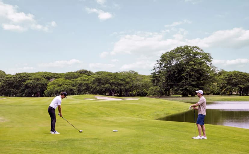 Two golfers on a Buenaventura fairway beside a lake, one playing a shot, with a large sculpted bunker guarding the green behind.