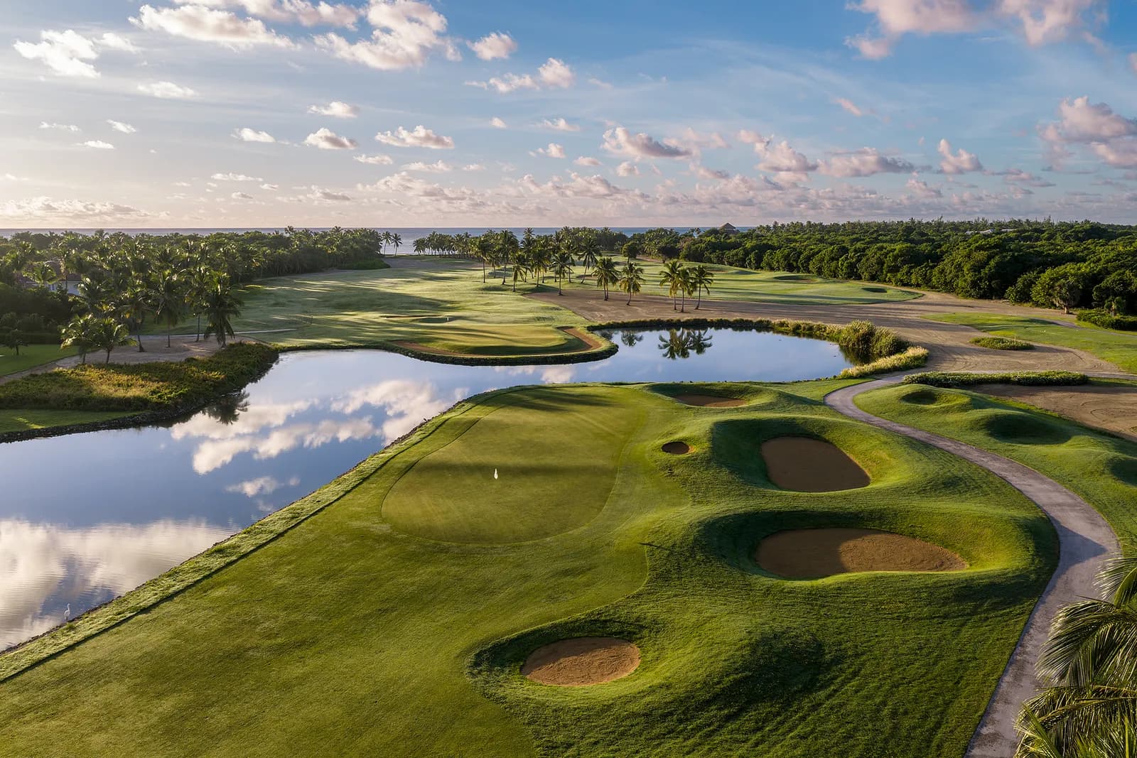 A La Cana green ringed by bunkers beside a calm pond reflecting clouds, with palms and the ocean in the distance