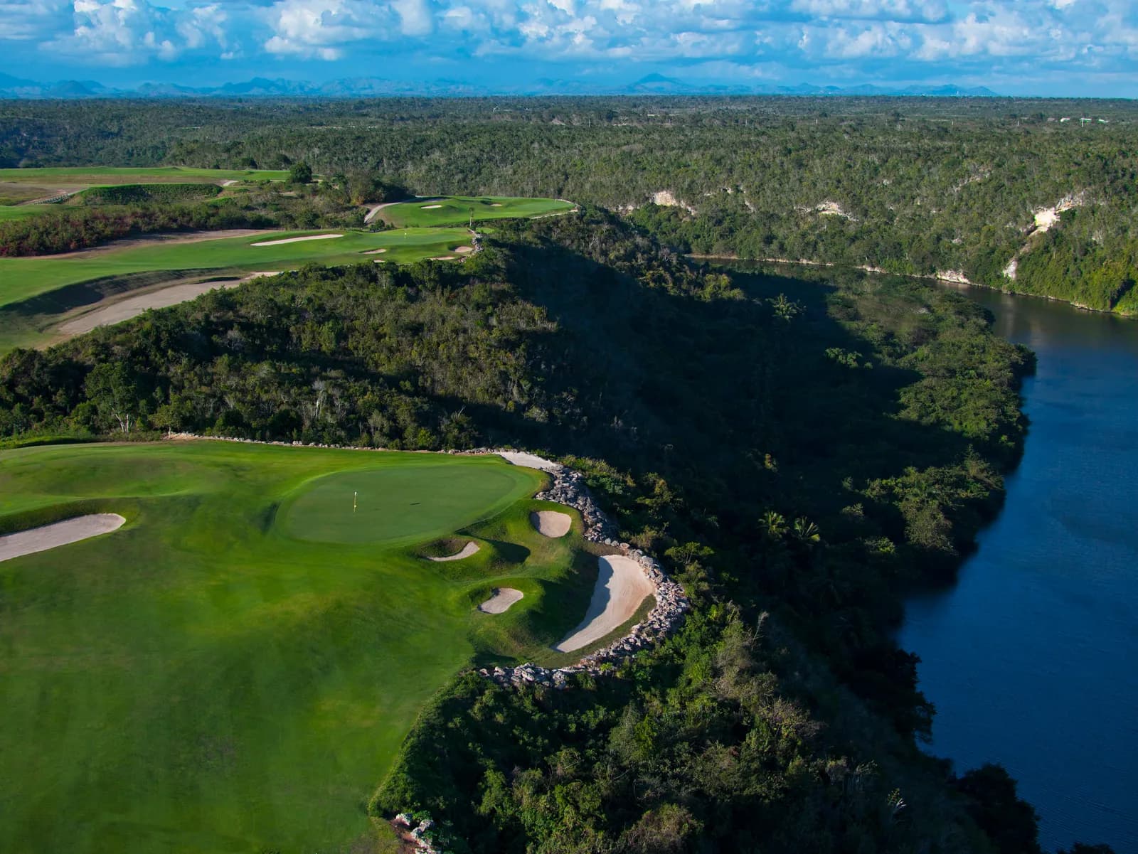 Aerial of a Dye Fore green and bunkers on a bluff high above the dark Chavón river, mountains on the horizon.
