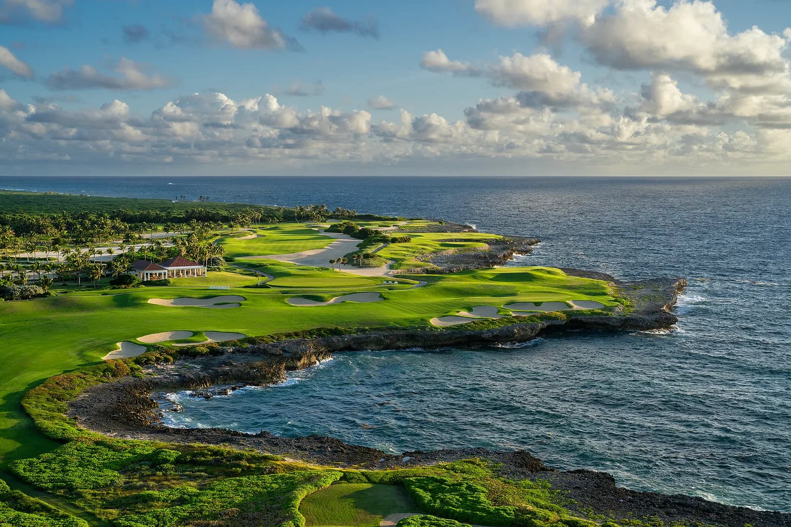 The 18th at Corales curving around a rocky cove at sunset, with the clubhouse above the cliff and a golden sky