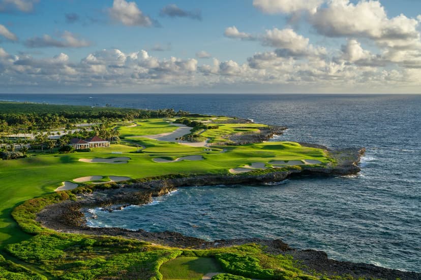 The 18th at Corales curving around a rocky cove at sunset, with the clubhouse above the cliff and a golden sky