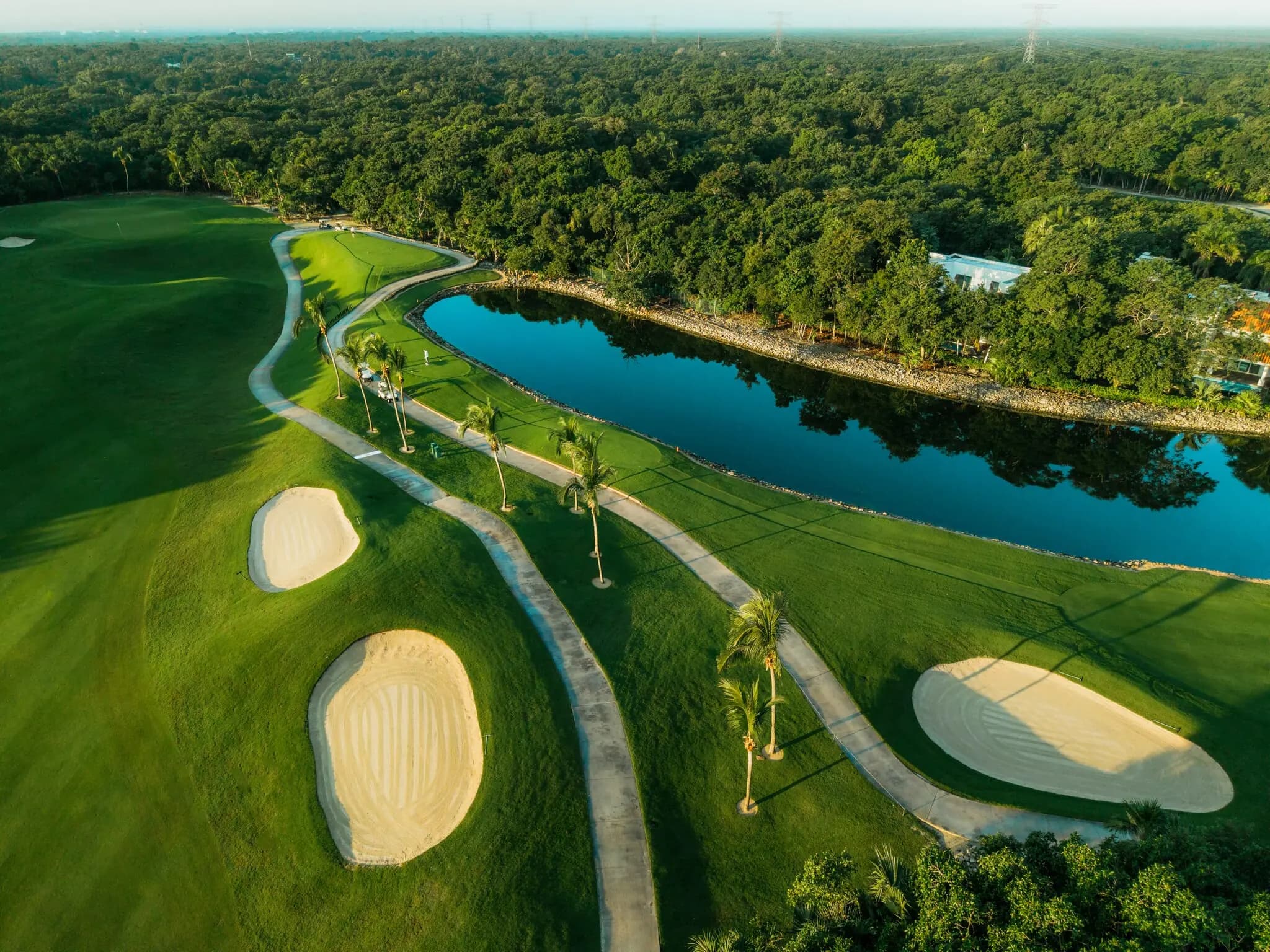 Golden-hour aerial of bunkers and a palm-lined cart path curving beside a lake at Iberostar Playa Paraiso Golf Club