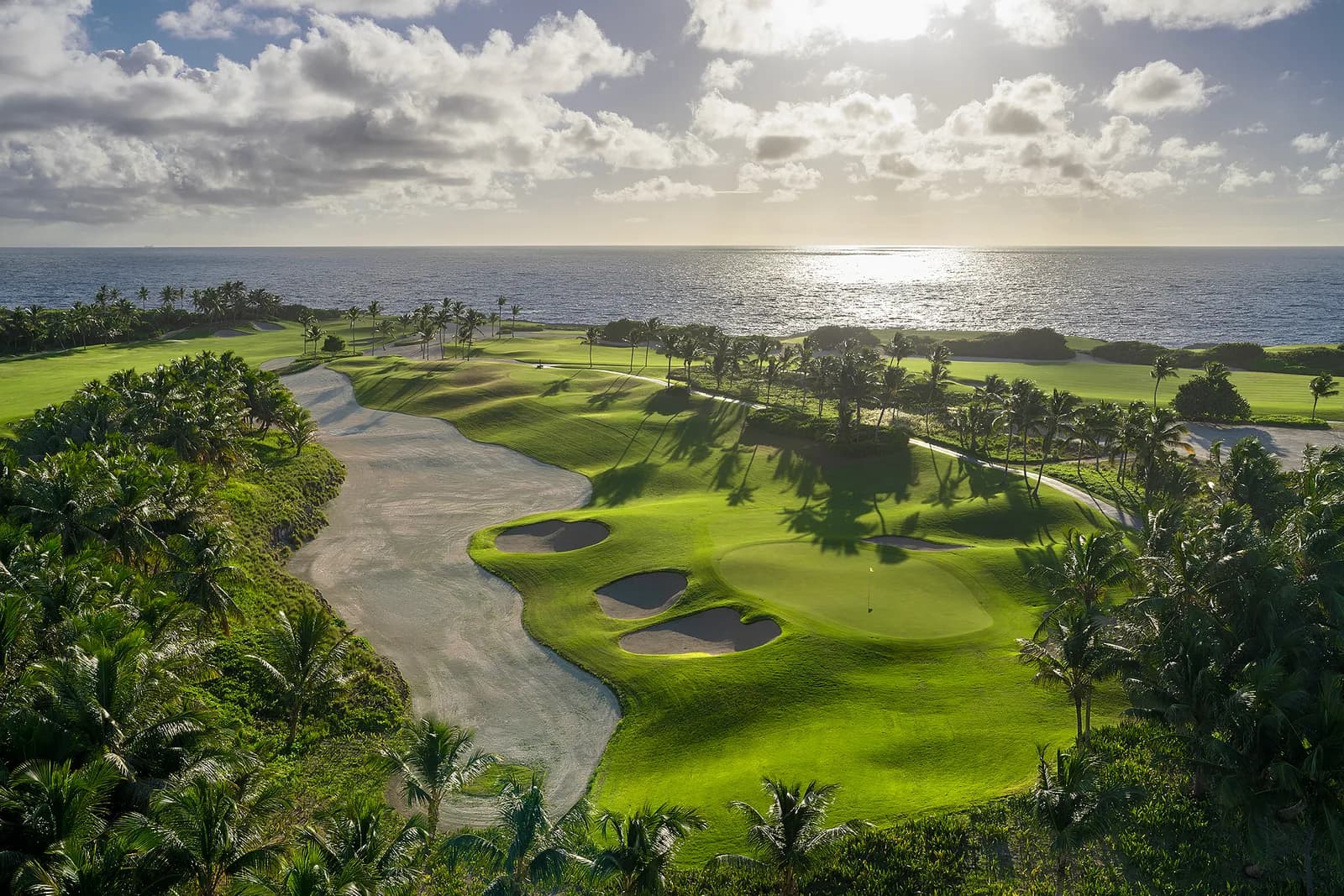 A waste sand lined Corales par 3 with palms casting shadows and bright ocean glare behind the green