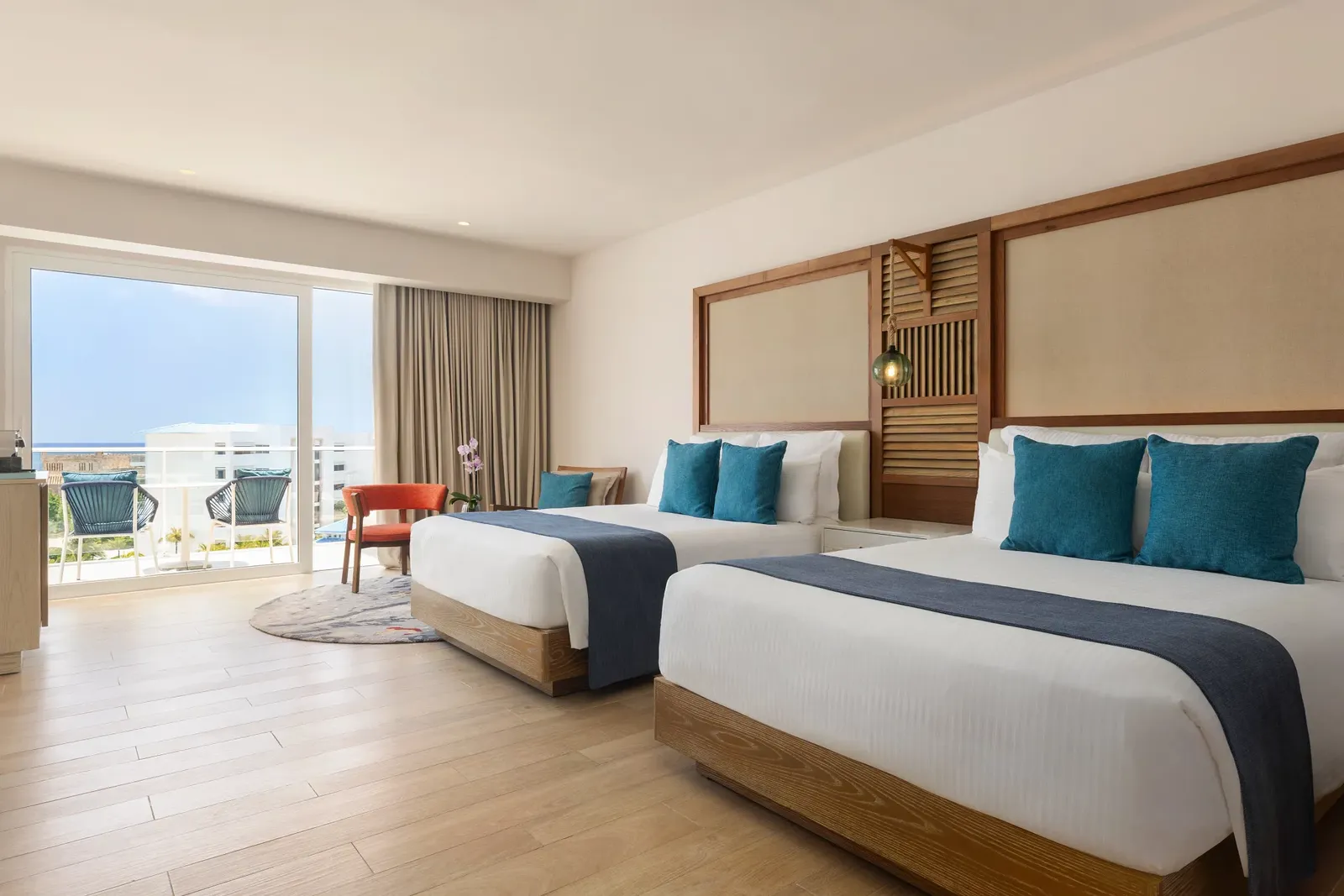 Guest room at Dreams Cap Cana with two beds, teal pillows, wood floors, and a balcony overlooking the resort toward the sea.