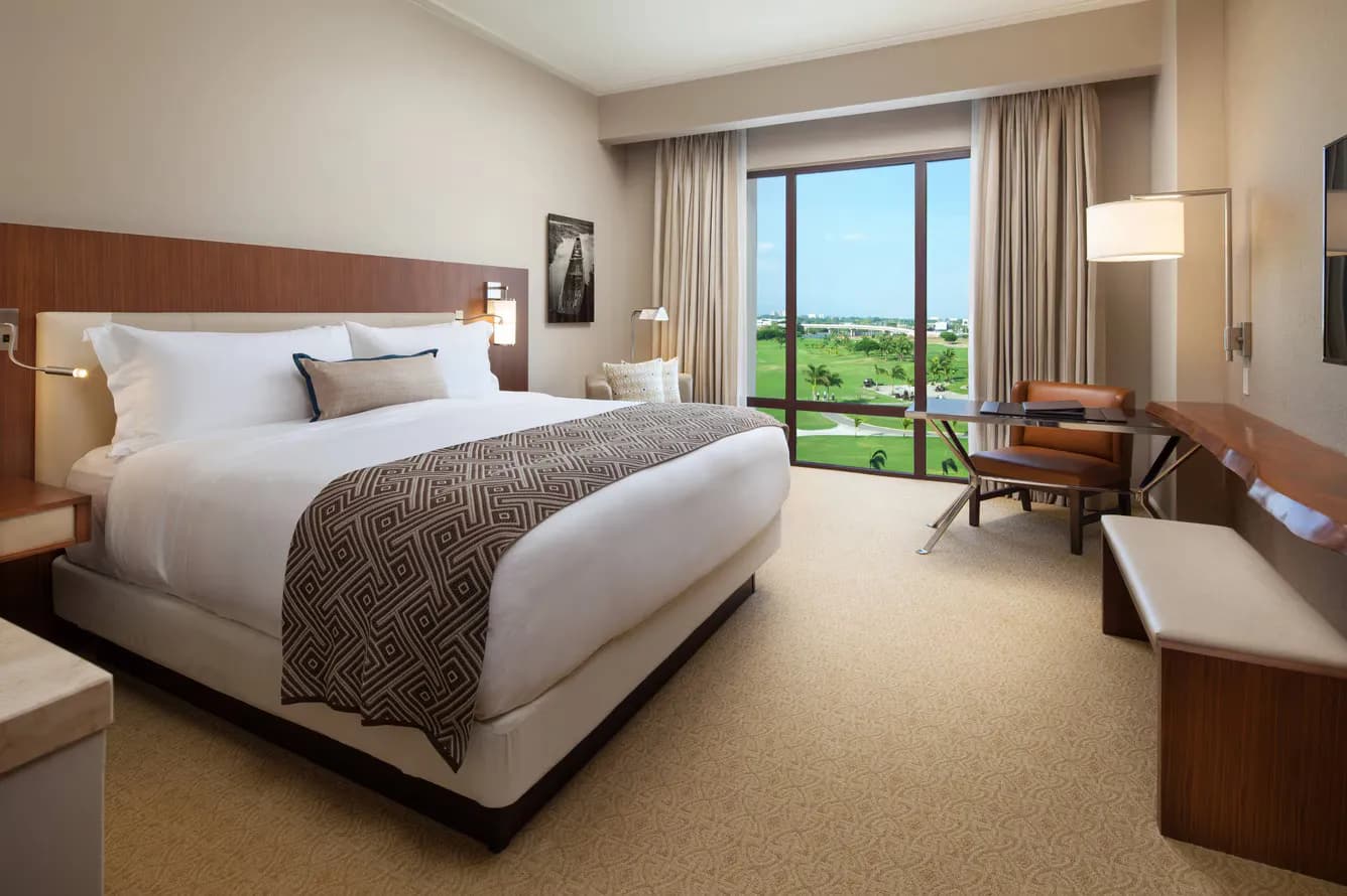 Hotel room with a king bed, patterned throw, desk with leather chair, and a floor-to-ceiling window overlooking green fairways and palms.