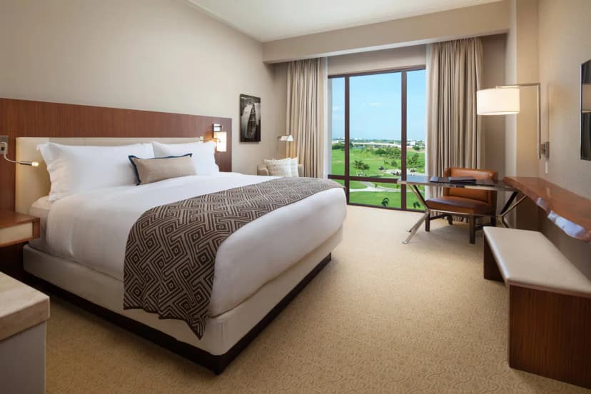 Hotel room with a king bed, patterned throw, desk with leather chair, and a floor-to-ceiling window overlooking green fairways and palms.