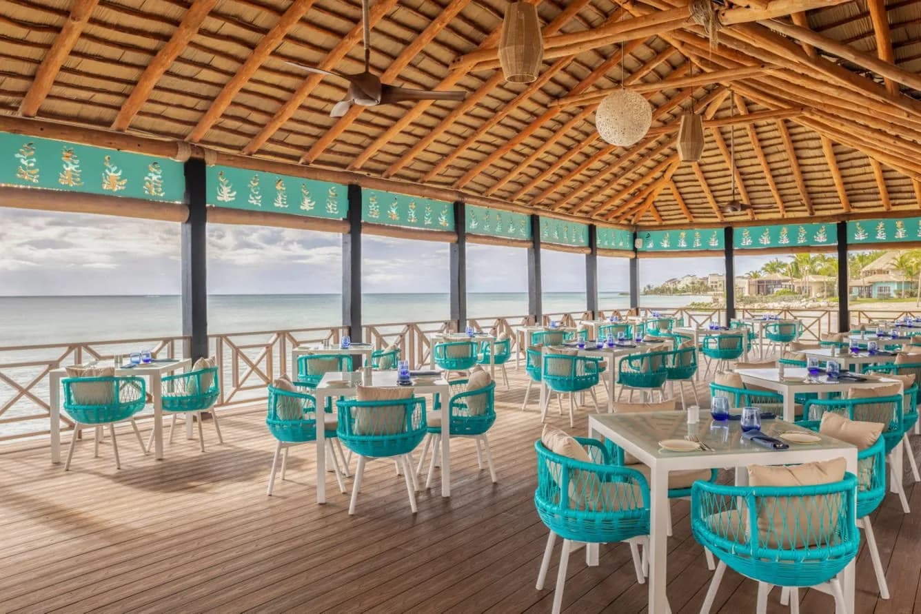 Open air overwater restaurant at Sanctuary Cap Cana with turquoise woven chairs, a thatched roof and sea views on three sides