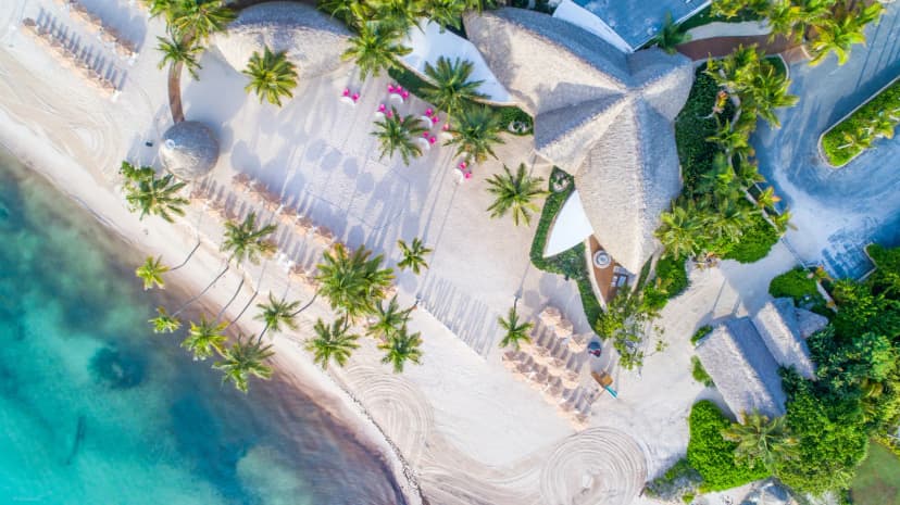 Top-down drone view of Playa Blanca restaurant's star-shaped thatched roof on white sand, pink chairs on the terrace, umbrellas, palms, and turquoise water.
