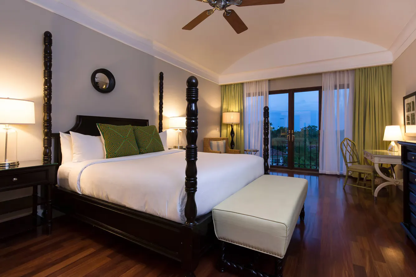 Guestroom with a dark-wood four-poster king bed, green Panamanian mola cushions, hardwood floors, and balcony doors open to dusk light.