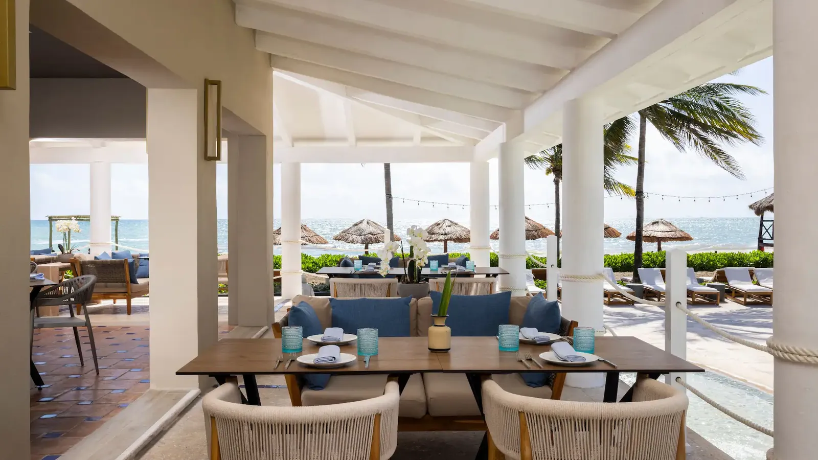 Open-air beachfront restaurant terrace with woven armchairs, set wooden tables, and thatched beach umbrellas along the sea behind.