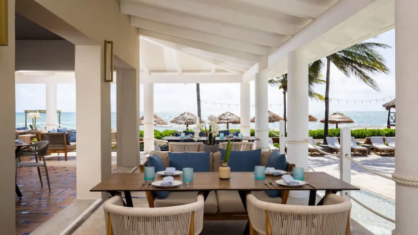 Open-air beachfront restaurant terrace with woven armchairs, set wooden tables, and thatched beach umbrellas along the sea behind.