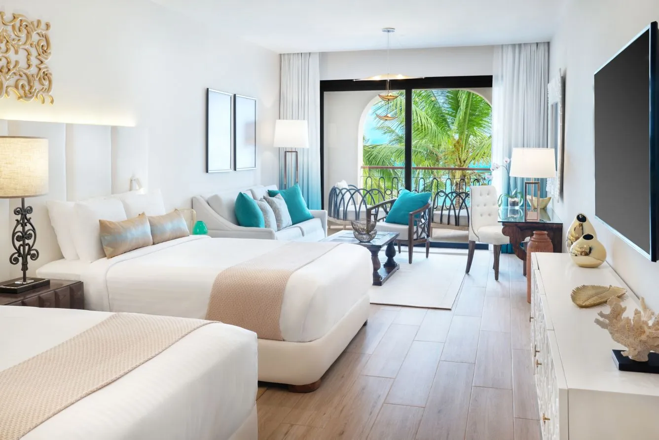 A guest room at Sanctuary Cap Cana with two beds, a sitting area, and an open balcony framing palm trees and the sea.