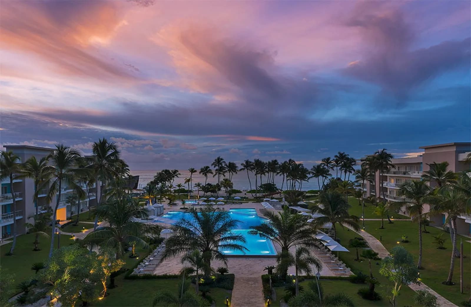 Evening view over the Westin Puntacana pool courtyard toward the sea, with a pink and violet sky above lit palms