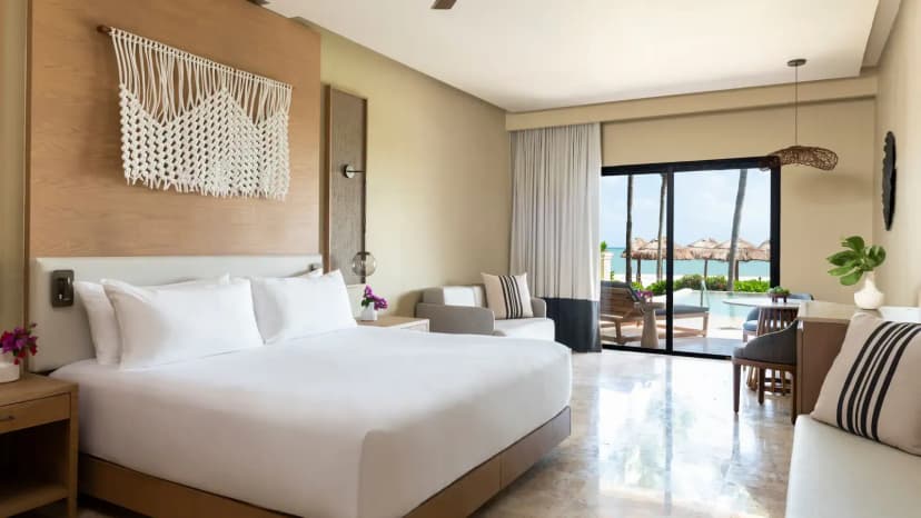 Suite interior with a king bed under macramé wall art, sitting area, and sliding glass doors opening to a terrace with palms and beach palapas beyond.