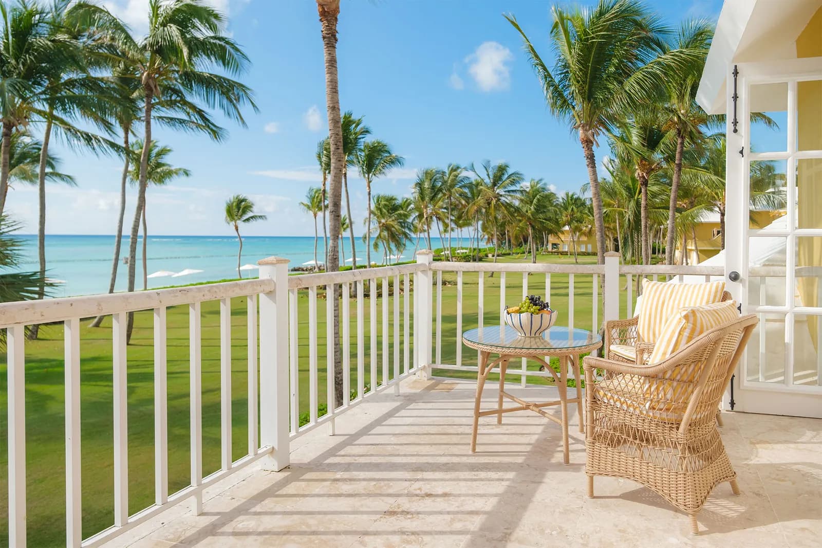 Private balcony of a Junior Suite Ocean View at Tortuga Bay with white railings, wicker furniture, palms, and the turquoise sea beyond.