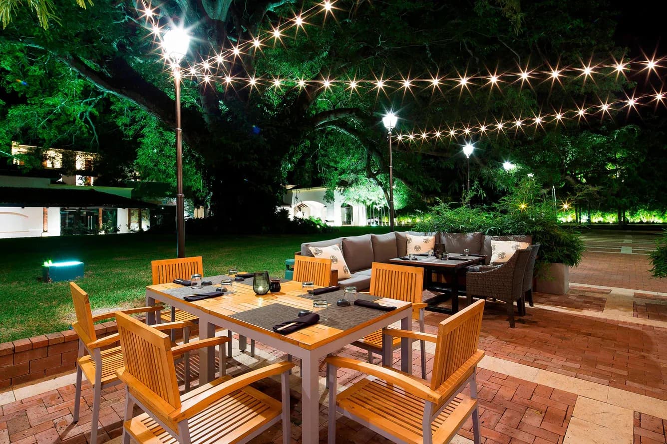 Outdoor evening dining terrace at Buenaventura with string lights strung under a huge Corotú tree, set wooden tables and a lounge sofa on brick pavers.