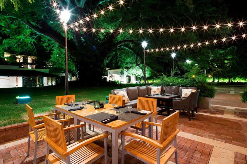 Outdoor evening dining terrace at Buenaventura with string lights strung under a huge Corotú tree, set wooden tables and a lounge sofa on brick pavers.