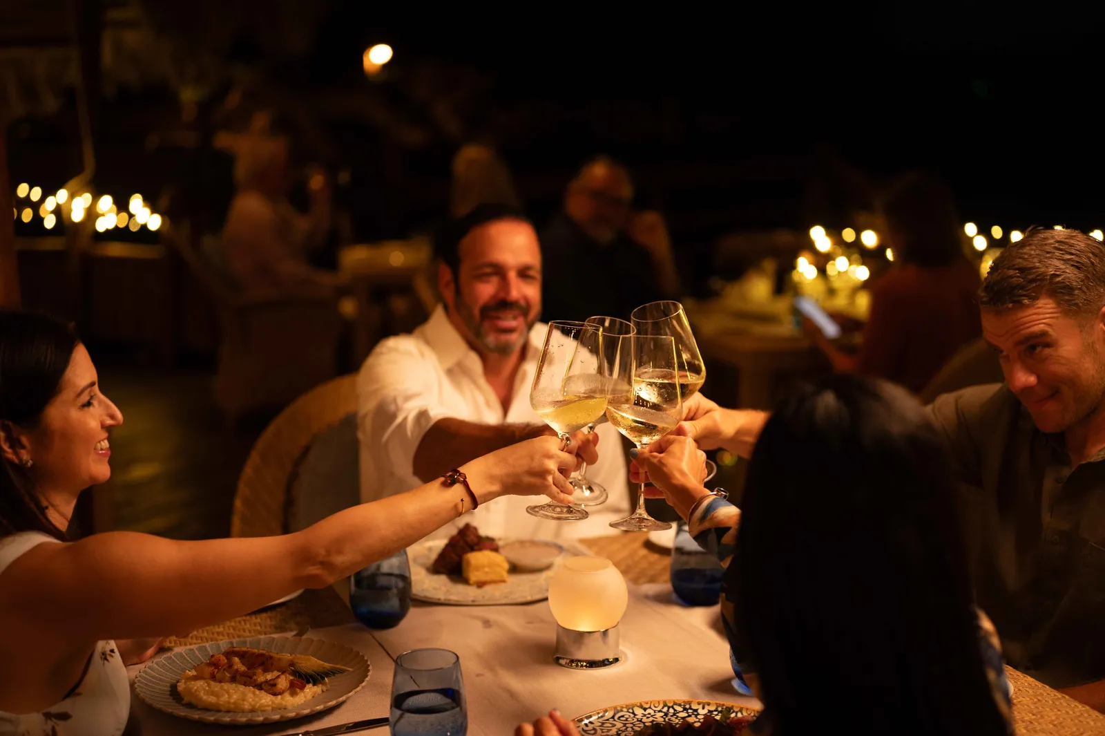 Diners raising wine glasses over a candlelit table at La Yola restaurant at night, string lights glowing behind them.
