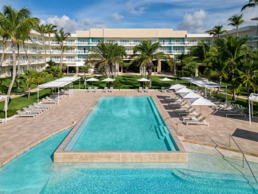 Aerial view of the main pool at The Westin Puntacana Resort, lined with white loungers and umbrellas, with the resort building and palms behind.