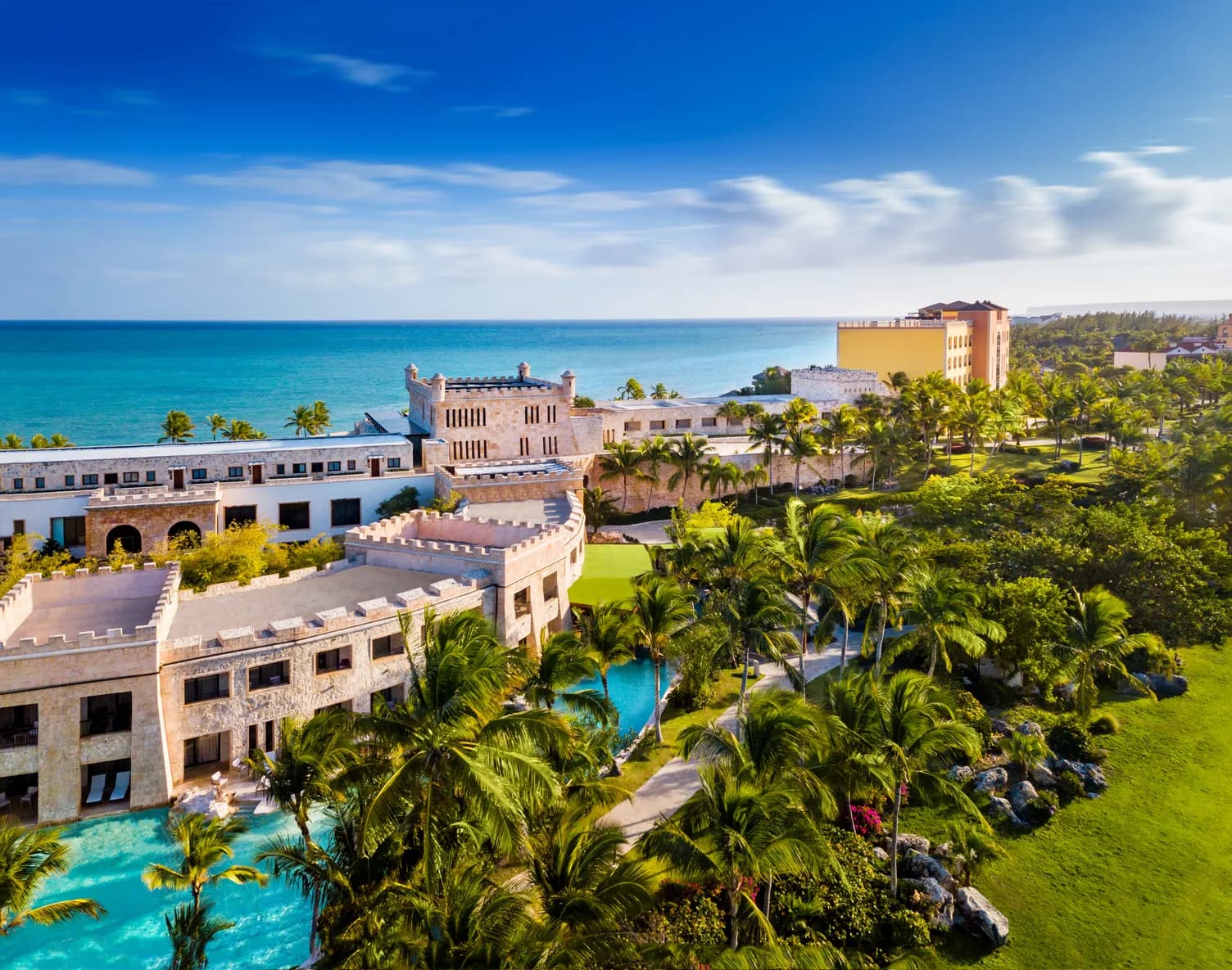 Aerial of Sanctuary Cap Cana's stone castle buildings, winding turquoise pools and palm gardens beside the sea