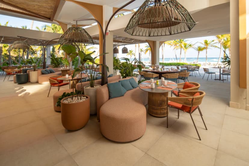 An open-air beachfront lounge at The Westin Puntacana with woven rattan pendant lights, cushioned seating and dining tables, looking out to palms and the sea.