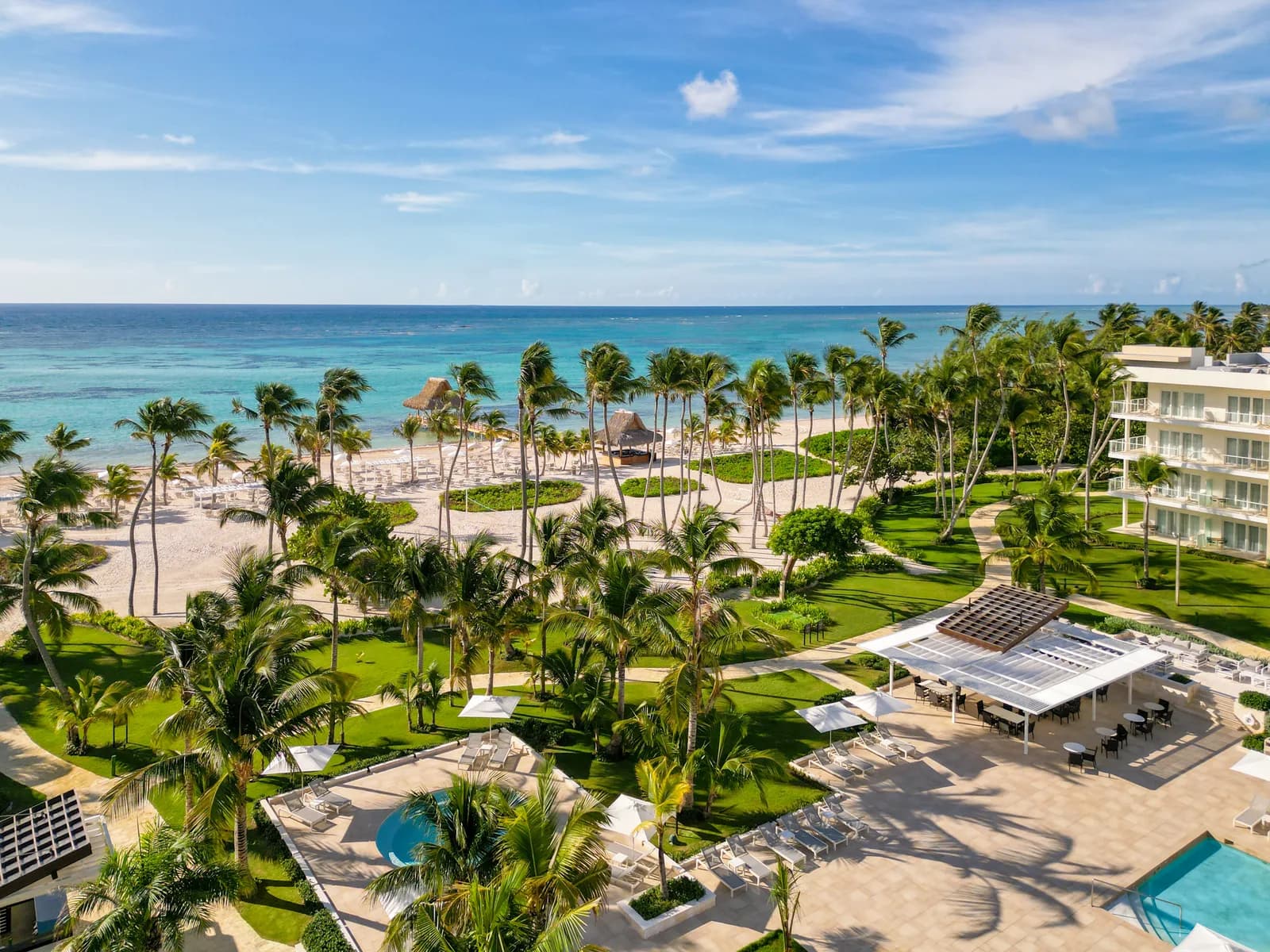 Aerial of The Westin Puntacana's beachfront: turquoise reef-protected water, white sand with palapas and loungers, palms, and a modern white hotel wing.