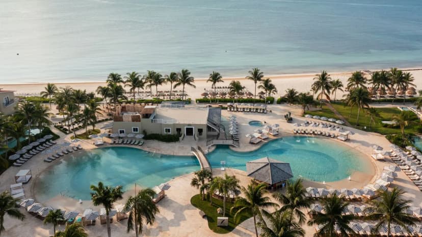 Aerial view of Hyatt Zilara Riviera Maya's two free-form pools with striped umbrellas, daybeds, and palms, the white-sand beach and turquoise sea beyond.