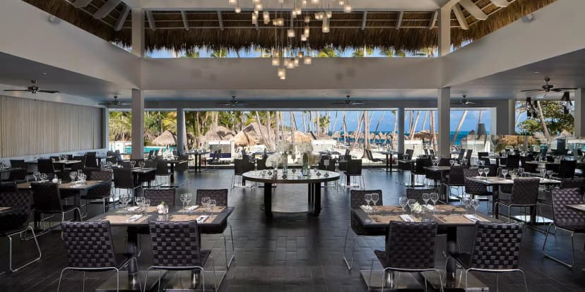 Open-air restaurant at Melia Caribe Beach with dark rattan chairs, chandeliers under a thatched roof, and palms and ocean beyond.