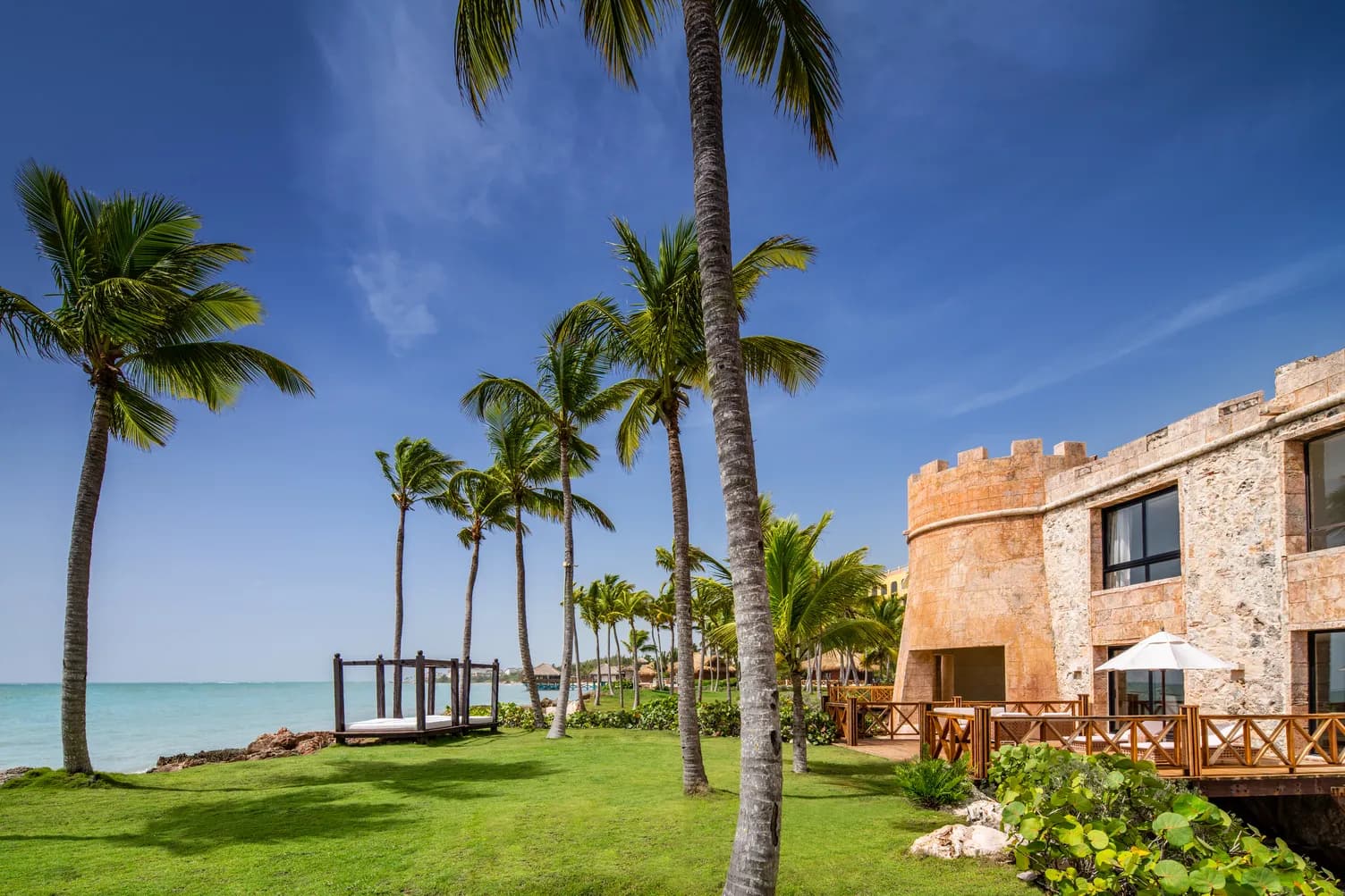 Oceanfront lawn at Sanctuary Cap Cana with tall palms, a wooden daybed and the stone castle tower at the water's edge