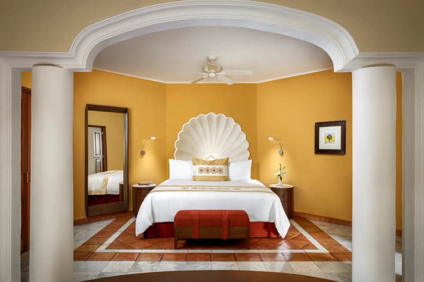 A suite bedroom at Casa Velas with a king bed under a carved shell headboard, ochre walls, a white archway and a terracotta tile floor.