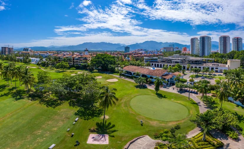 Aerial view of Marina Vallarta Golf Club showing the red-tiled clubhouse, a practice green and fairways, with the Puerto Vallarta skyline and mountains beyond.