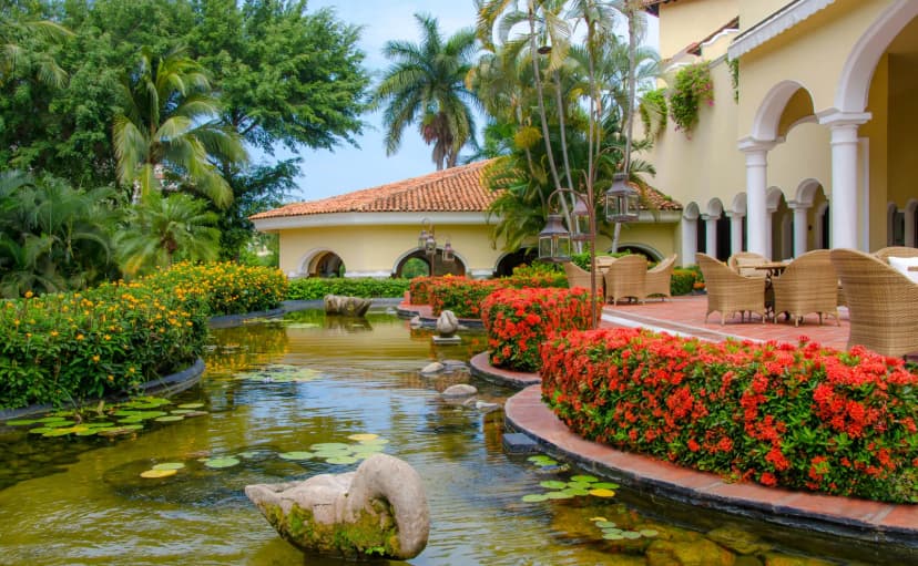 A garden courtyard at Casa Velas with a lily pond, red flowering hedges, a stone swan sculpture and a terrace of wicker chairs beneath tiled arches.