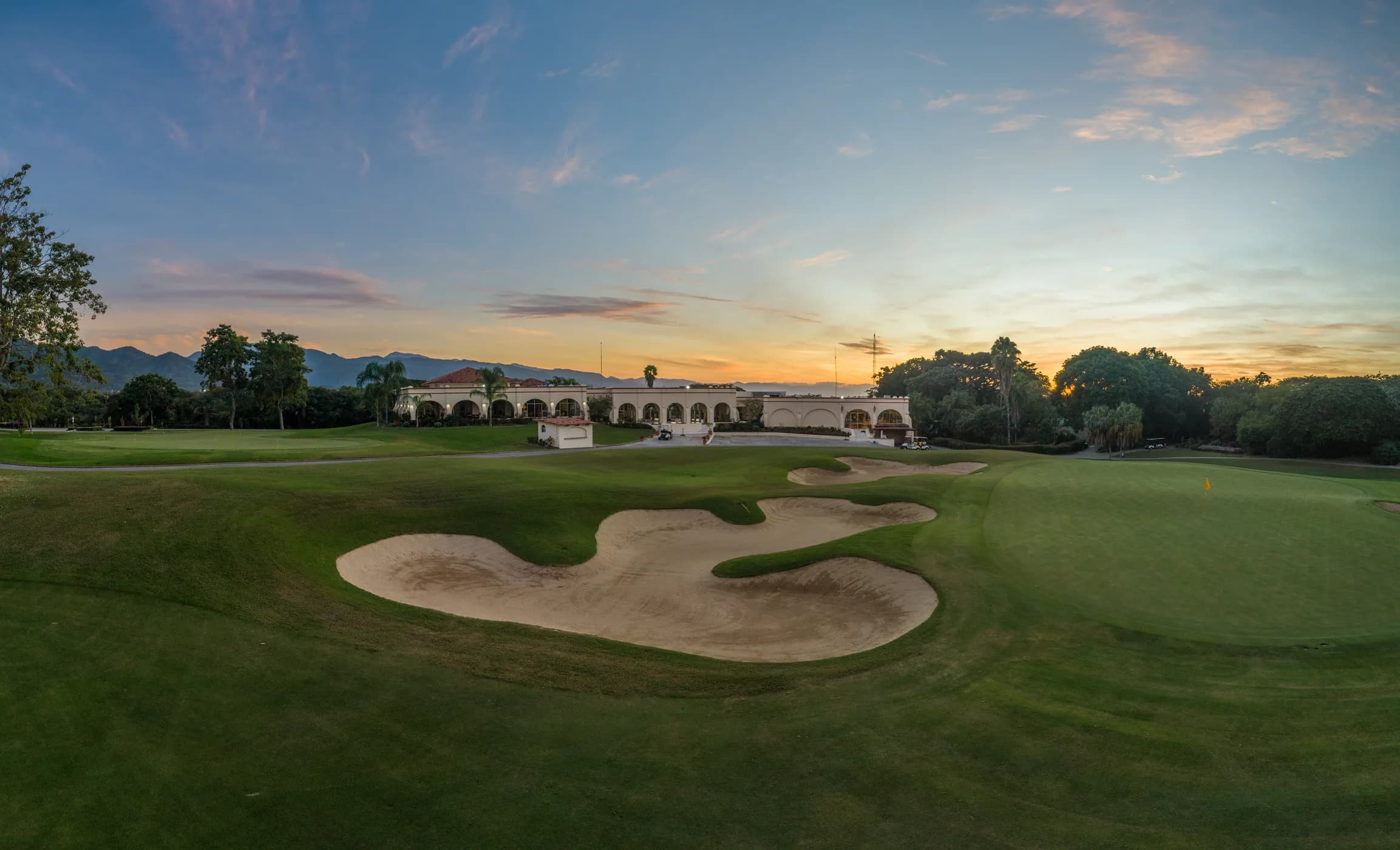 Sand bunkers before a green at sunset, with the arched Marina Vallarta clubhouse and the Sierra Madre mountains on the horizon.